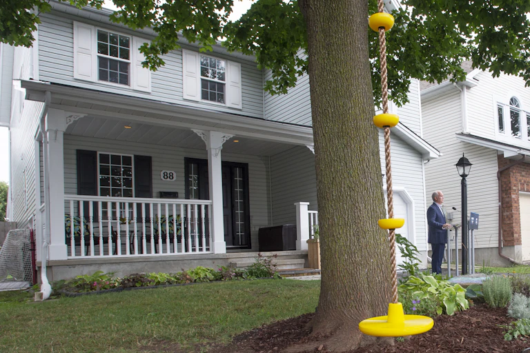A tree with a yellow swing is positioned infront of a light blue house which has a big white front porch. Erin O'Toole stands on the far right corner.