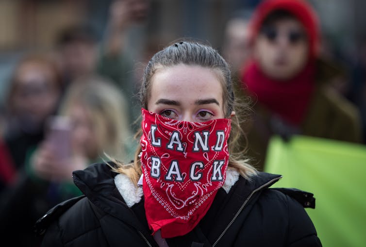 A woman wears a bandana over her face reading 'Land Back'