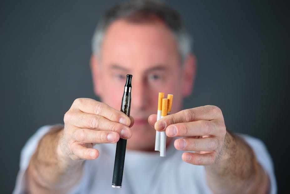 Older man holds up an e-cigarette in one hand, and three cigarettes in the other.