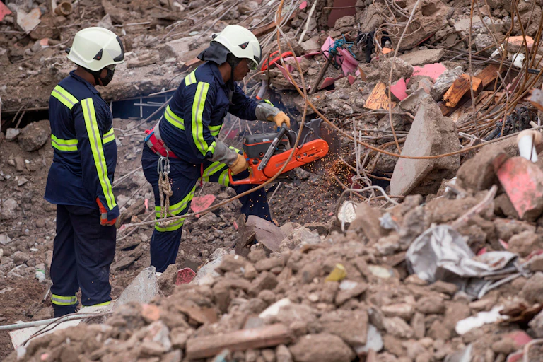 Two rescue workers cut through metal cables amid earthquake rubble.