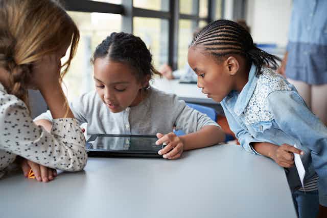 Three young girls look at an iPad  during class.
