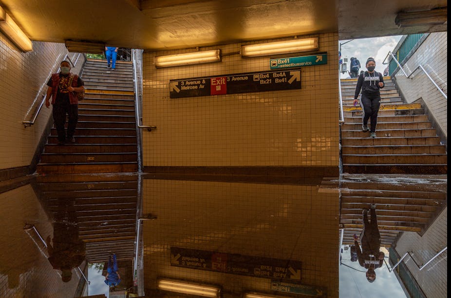 Commuters walk into a flooded subway station. The floor is covered with standing water.