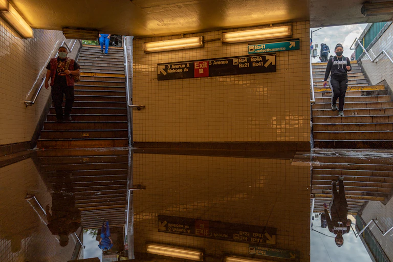 Commuters walk into a flooded subway station. The floor is covered with standing water.