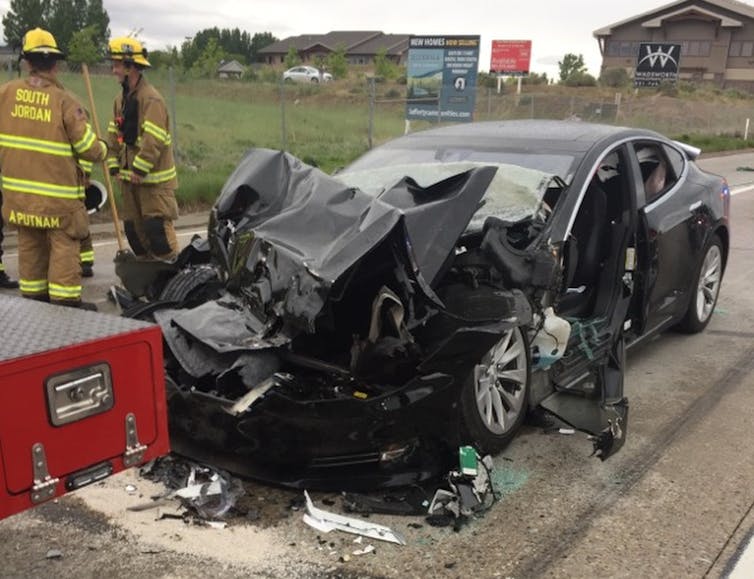 Two firefighters stand beside a wrecked car with a crumpled front end