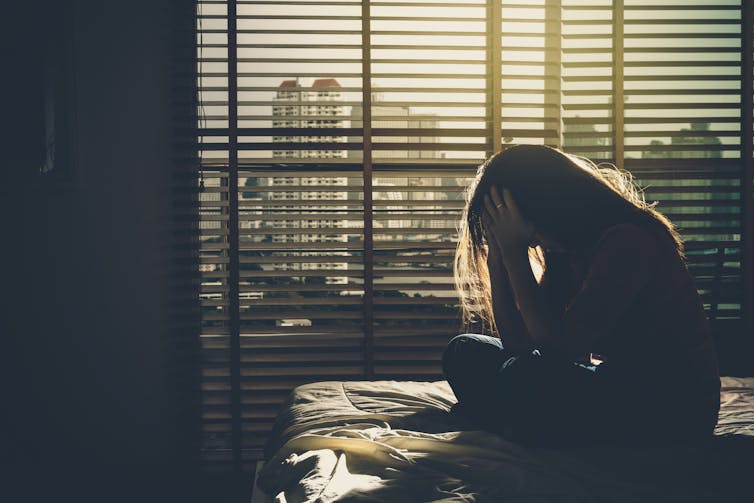 A woman sits on a bed in the dark, appears depressed.