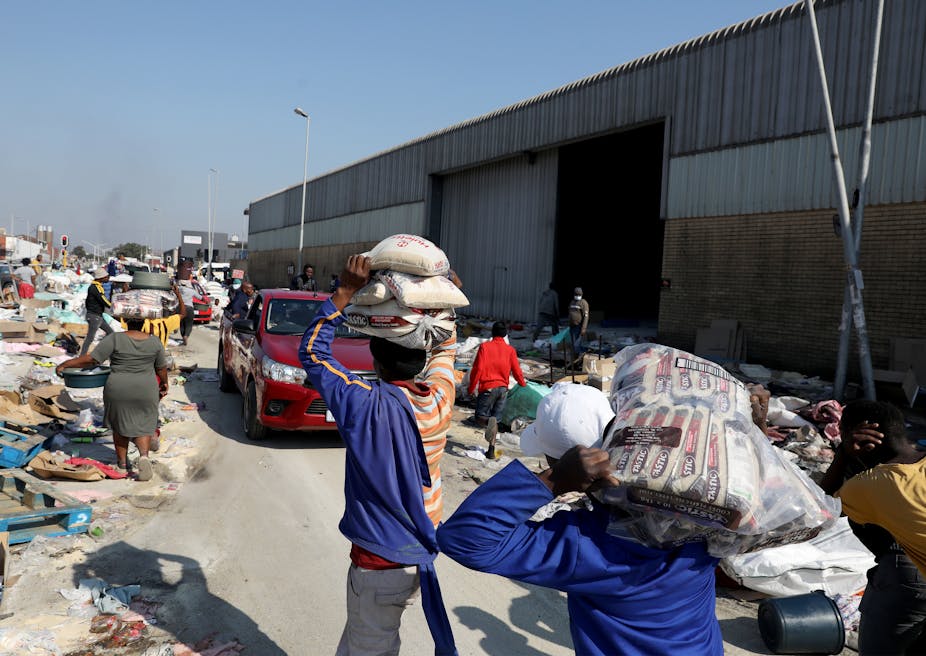 Men and women carry piles of groceries from a warehouse on their heads. 