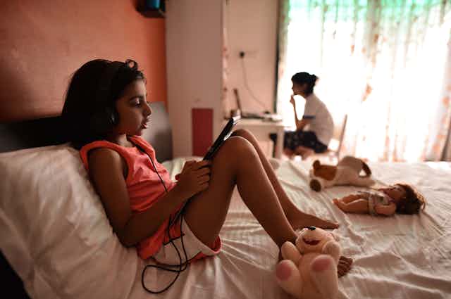 Girl wearing headphones works on a tablet while sitting on a bed