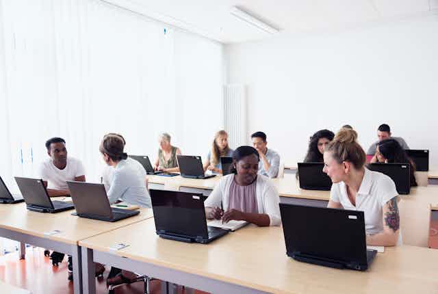 A group of people in a classroom talk among themselves while having their laptops open.