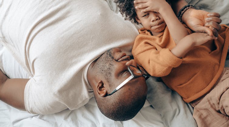 A father and child talk lying down and the boy looks a little apprehensive while the father holds his hand.