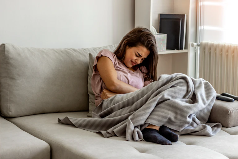 A woman sitting on her couch wrapped in a blanket, holding her stomach in pain because of her endometriosis.