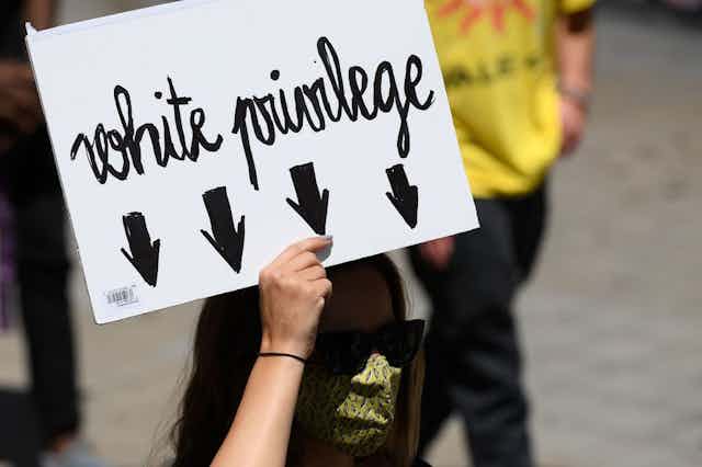A woman in a green face mask and shades holds a black and white placard reading 'White Privilege' with arrows pointing downward to indicate the woman holding the poster
