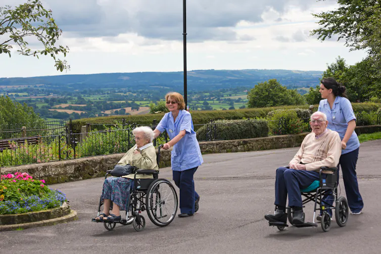 Two old people being pushed outside in wheelchairs by carers