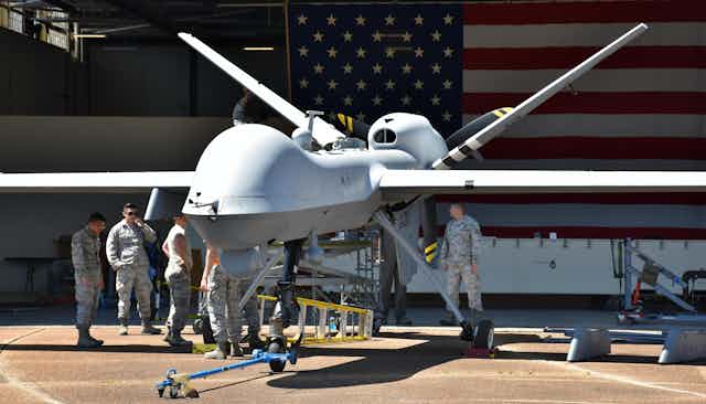 US military personnel performing maintenance on a drone in an airplane hangar, with a large American flag in the background