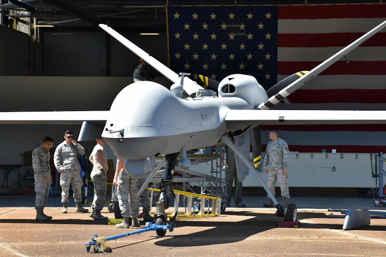 US military personnel performing maintenance on a drone in an airplane hangar, with a large American flag in the background