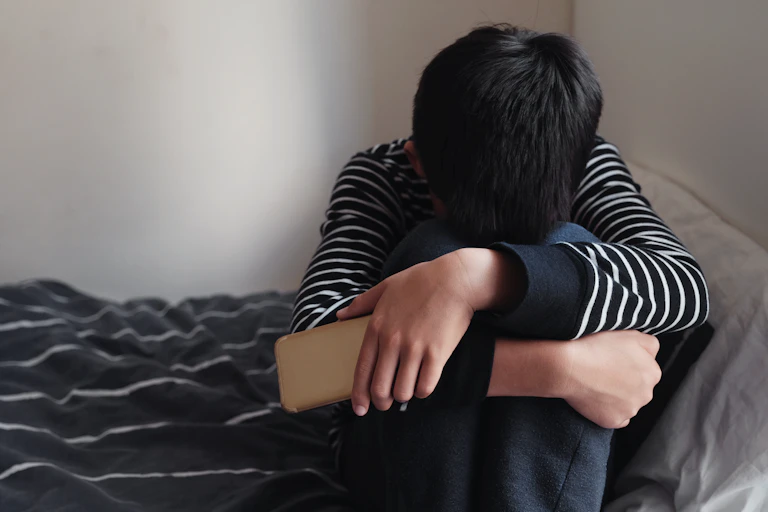 Boy on the bed with knees up and head in his knees, holding mobile phone in one hand.