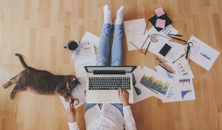 Person sitting on floor at home with laptop, paperwork and cat.