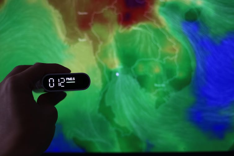 A hand holds a sensor depicting PM2.5 levels in front of a world map.