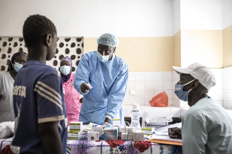 Médecin en blouse bleue parle à un patient dans une salle de consultation, avec une table couverte de médicaments entre eux