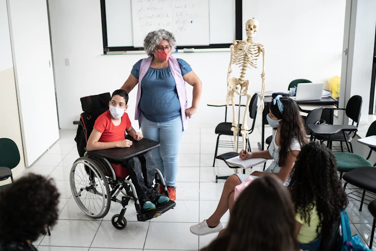A teacher stands beside student using a wheelchair while other students sit in desks