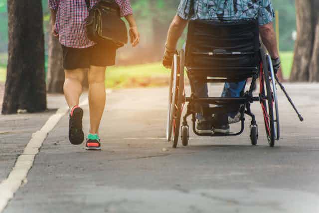 Woman walks next to man in a wheelchair, down a path in a park the sun sets ahead