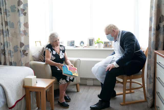 Boris Johnson talking to a resident in a care home. He is wearing a face mask and plastic apron and she is showing him a book.