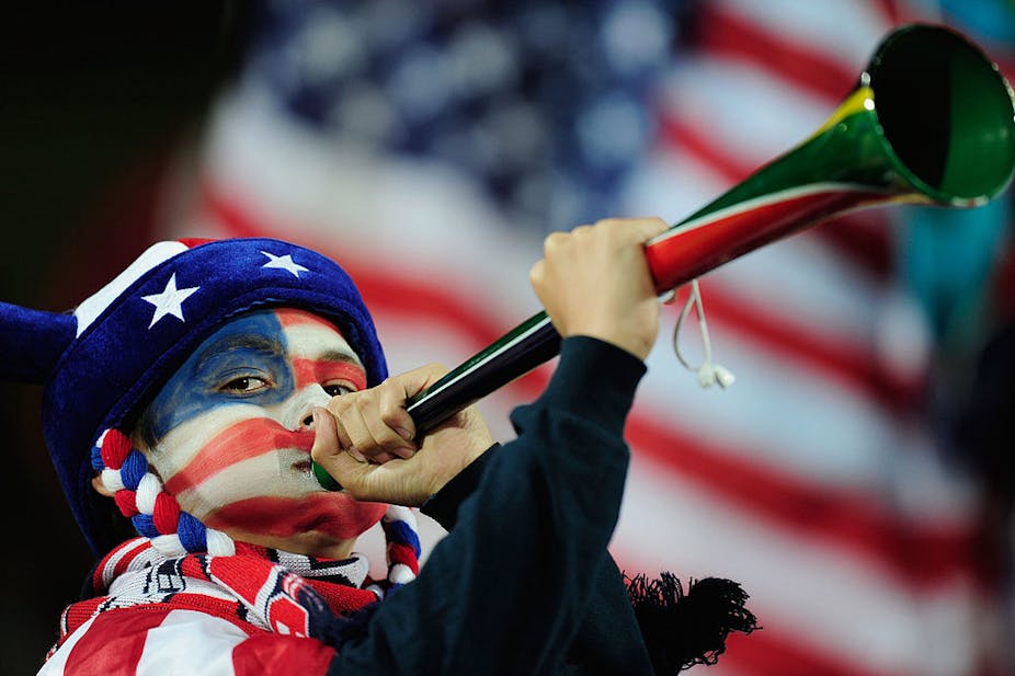 Boy with red, white and blue face paint and wearing a hat with stars on it blows a trumpet-like instrument; US flag in background