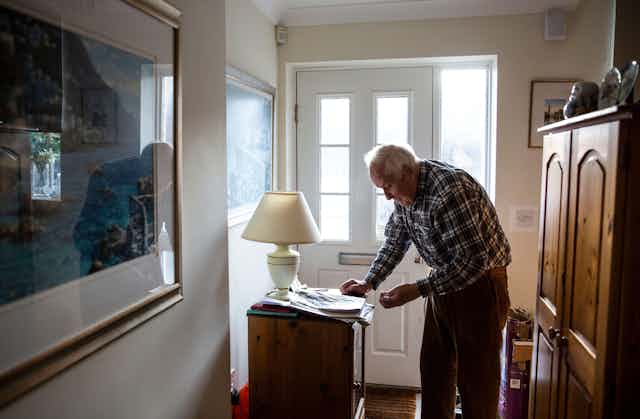 An elderly man stands in his hallway at home, reading a newspaper