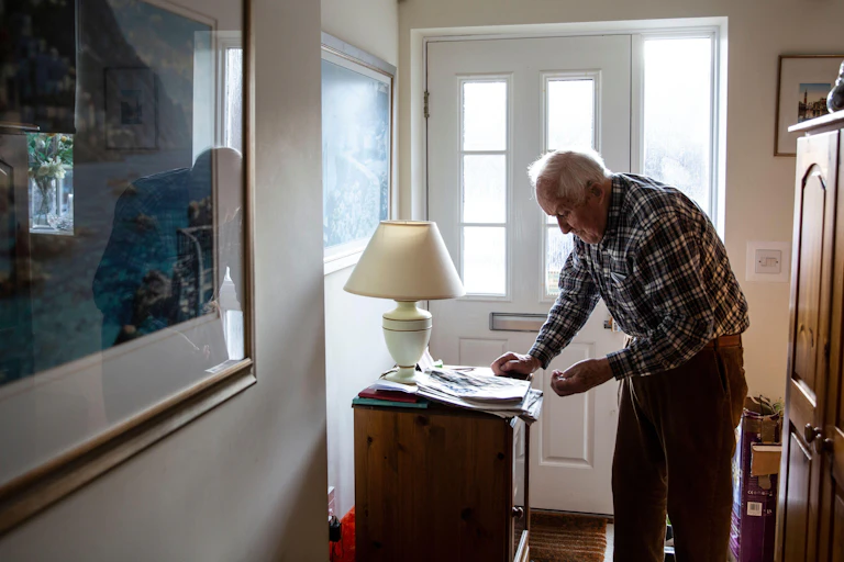 An elderly man stands in his hallway at home, reading a newspaper