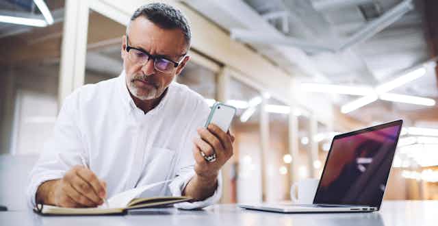 Un homme prend des notes dans un calepin, son cellulaire à la main, à côté d'un laptop ouvert