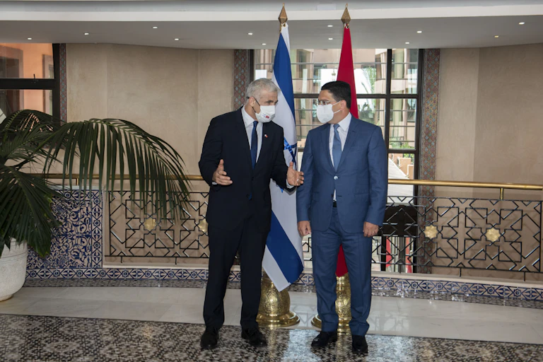 Two men wearing suits and COVID-19 masks chat with their backs to the flag of Israel on the left, and that of Morocco to the right.