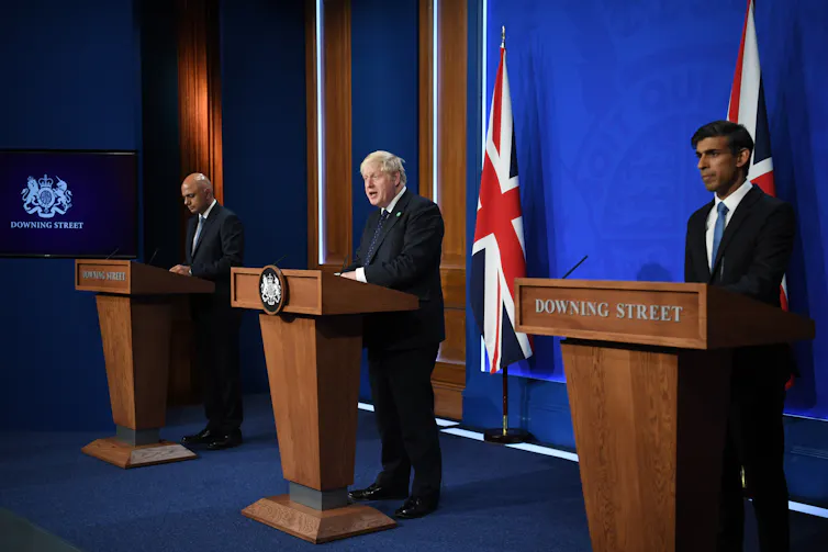 Health secretary Sajid Javid, Prime Minister Boris Johnson, and Chancellor Rishi Sunak behind podiums.