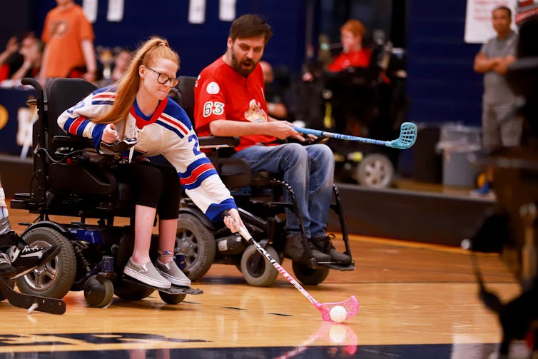 An athlete in a powered wheelchair leans forward to hit a ball with a hockey stick