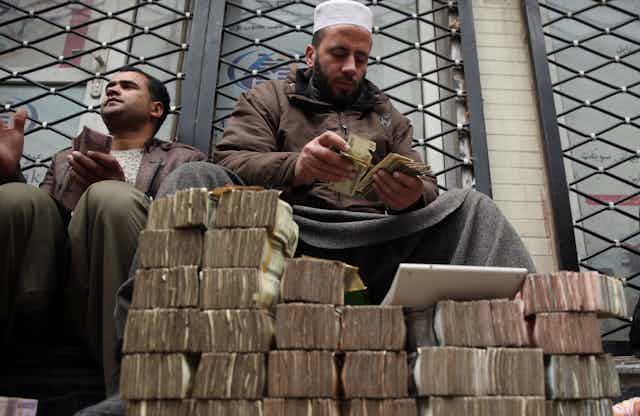 An Afghan money changer counts a pile of currency in Kabul as many piles of Aghani notes sit int he foreground