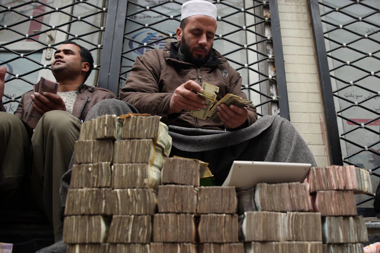 An Afghan money changer counts a pile of currency in Kabul as many piles of Aghani notes sit int he foreground