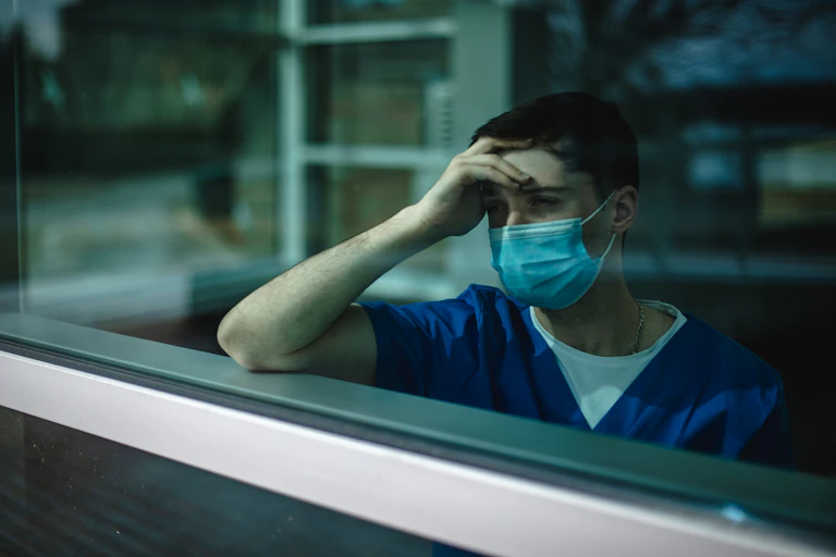 masked male nurse holding head and looking out window
