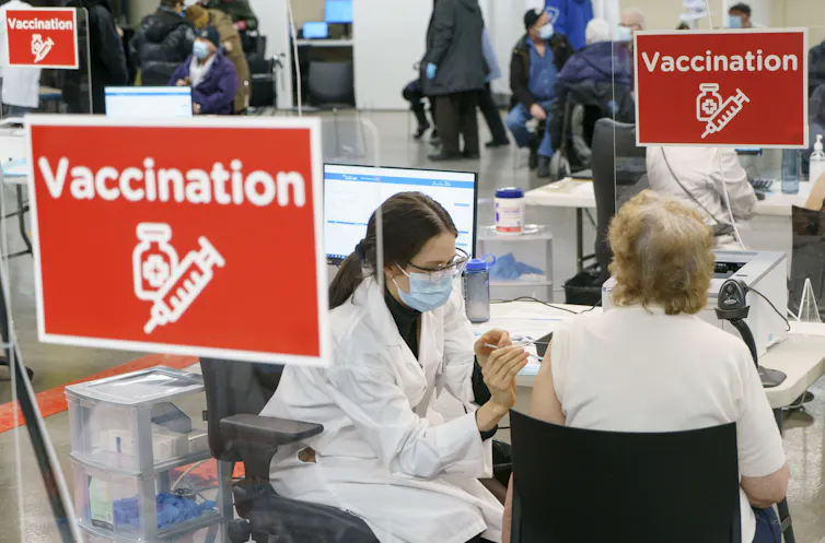 A woman administers a vaccine to another woman, seated, from behind