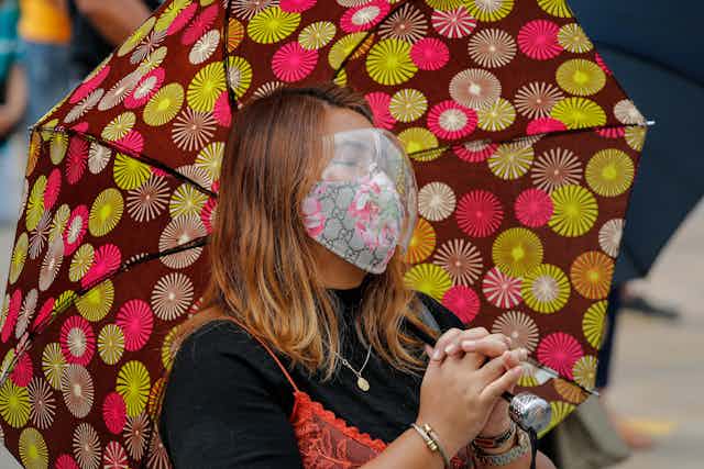 Philippino woman holding an umbrella and wearing a mask and goggles, closes her eyes and folds her hands, preying.
