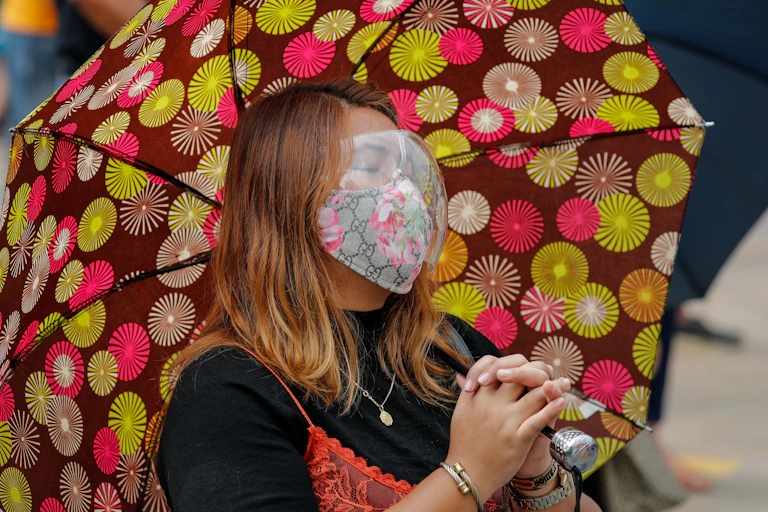Philippino woman holding an umbrella and wearing a mask and goggles, closes her eyes and folds her hands, preying.