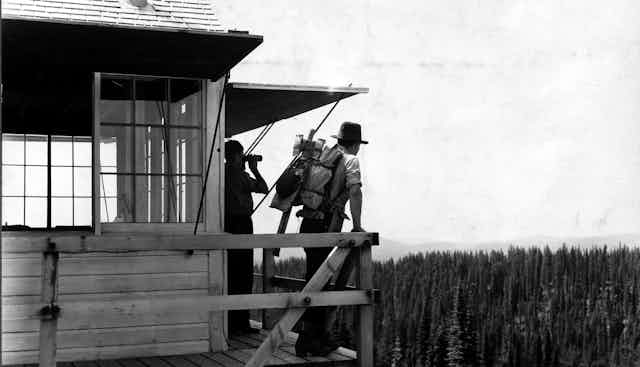 In an old photo, two people stand on a tall fire tower overlooking miles of pines trees.