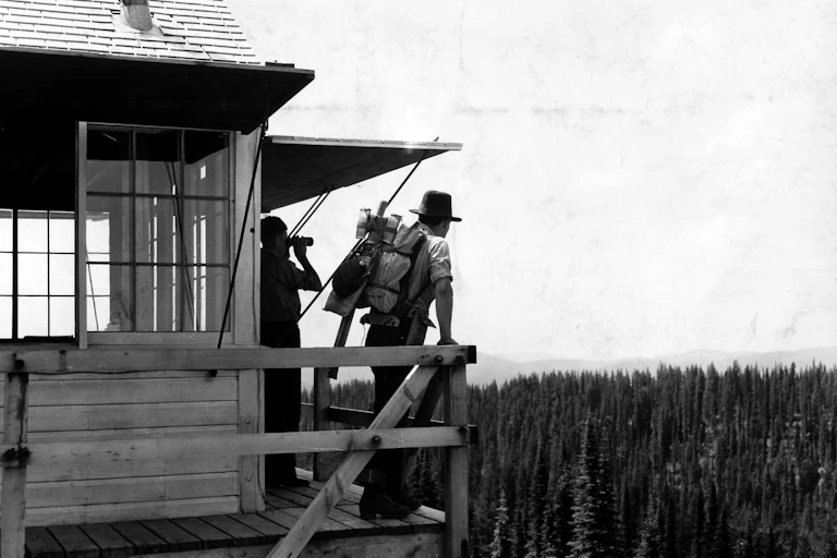 In an old photo, two people stand on a tall fire tower overlooking miles of pines trees.