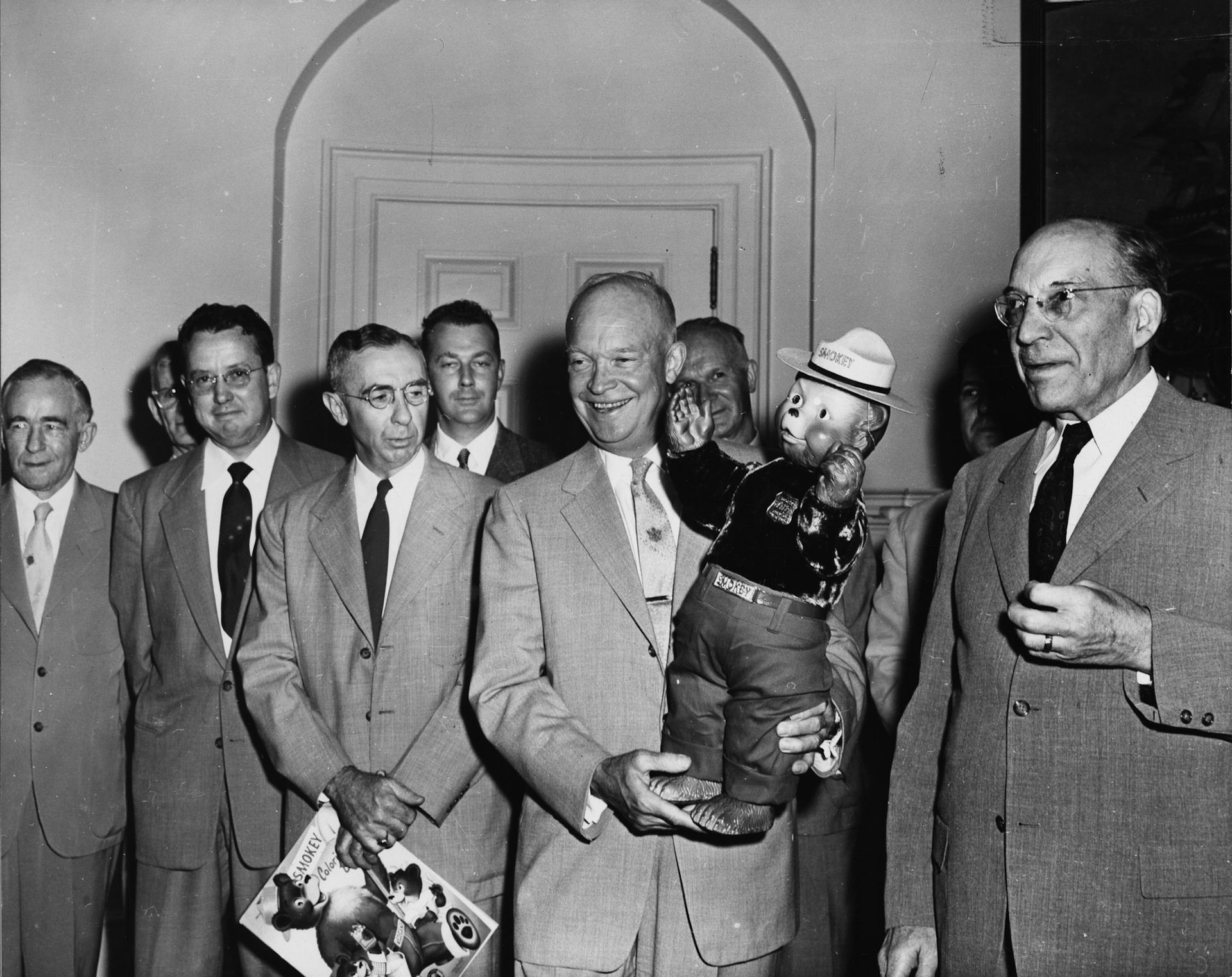 President Eisenhower smiles as he holds up the toddler-sized Smokey Bear doll with men in suits flanking him.