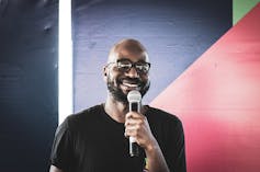 against a backdrop of angular colour block paintings, a bald bearded man in glasses smiles as he talks into a microphone.