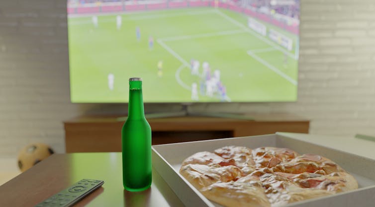 Bottle of beer and pizza on table in front of TV showing football game.