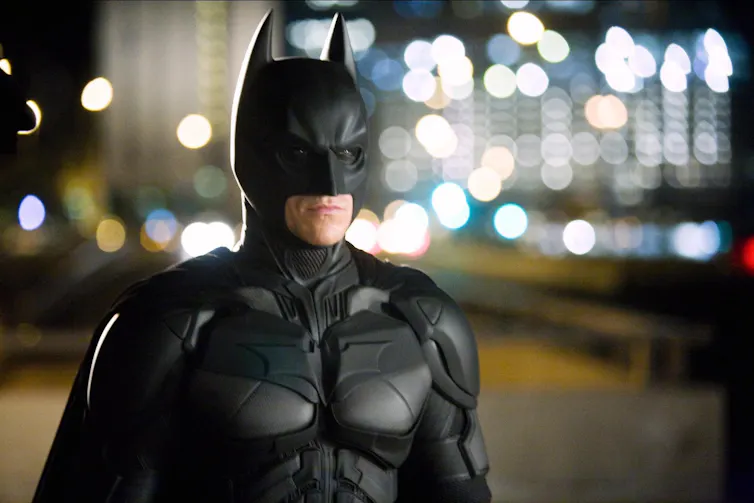 Batman stands in front of buildings.