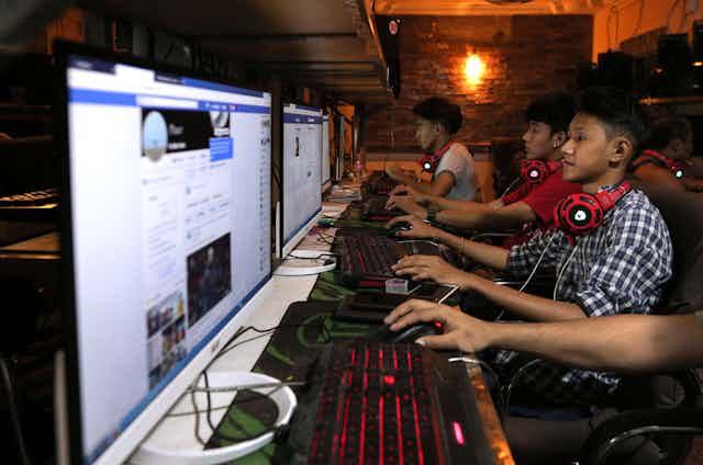 A group of young people sit in front of computer screens and keyboards that are lined up on a long table in a dark room