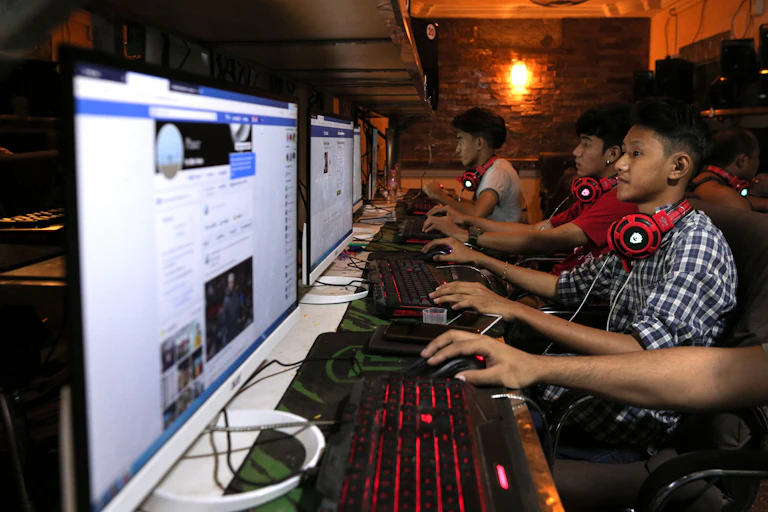 A group of young people sit in front of computer screens and keyboards that are lined up on a long table in a dark room