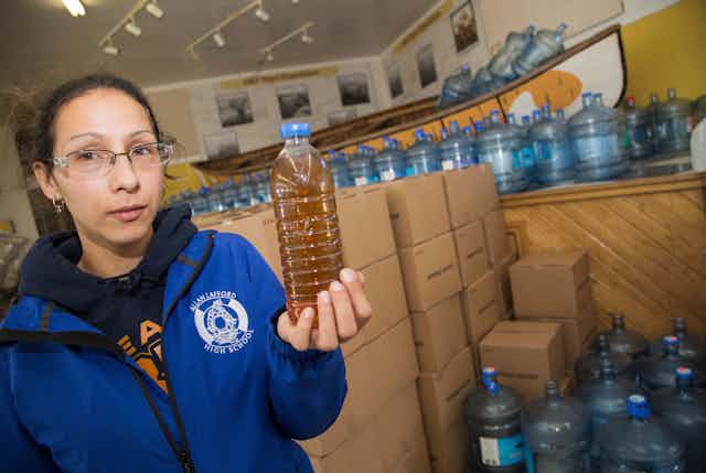 (Woman holds up a plastic water bottle full of dark brown water.