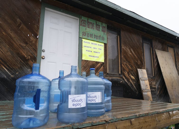 If All Parties Agree That We Need To End Drinking Water Advisories In Indigenous Communities, Why Haven'T We? 43 Water Bottles Sit On A Porch, Behind Reads A Sign That Says 'Grassy Narrows Water Supply' With A Smaller Sign That Reads 'Please Do Not Block Loading Area For Water Pickup'