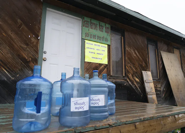 Water bottles sit on a porch, behind reads a sign that says 'Grassy Narrows Water Supply' with a smaller sign that reads 'please do not block loading area for water pickup'