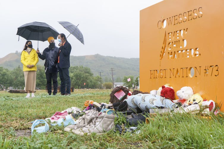 Three people stand holding umbrellas in the background, a sign in the front in orange reads 'ever child matters' teddy bears and other memorabilia lay on the ground in front of it